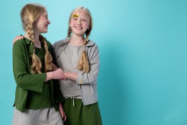 Twin blond girls with braids smiling and embracing against a turquoise background, indoors.